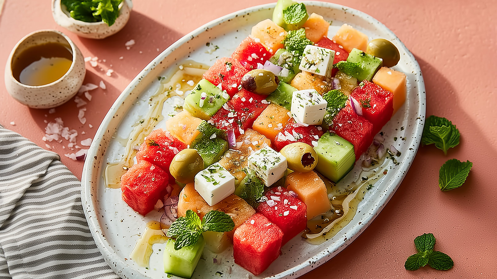 Ein Teller mit bunten Stückchen von Wassermelone, Honigmelone und grünem Obst. Darauf liegen einige Oliven, Feta-Würfel und frische Minzblätter. Das Gericht ist mit Olivenöl und Gewürzen garniert. Der Hintergrund ist in sanften, warmen Tönen gehalten.