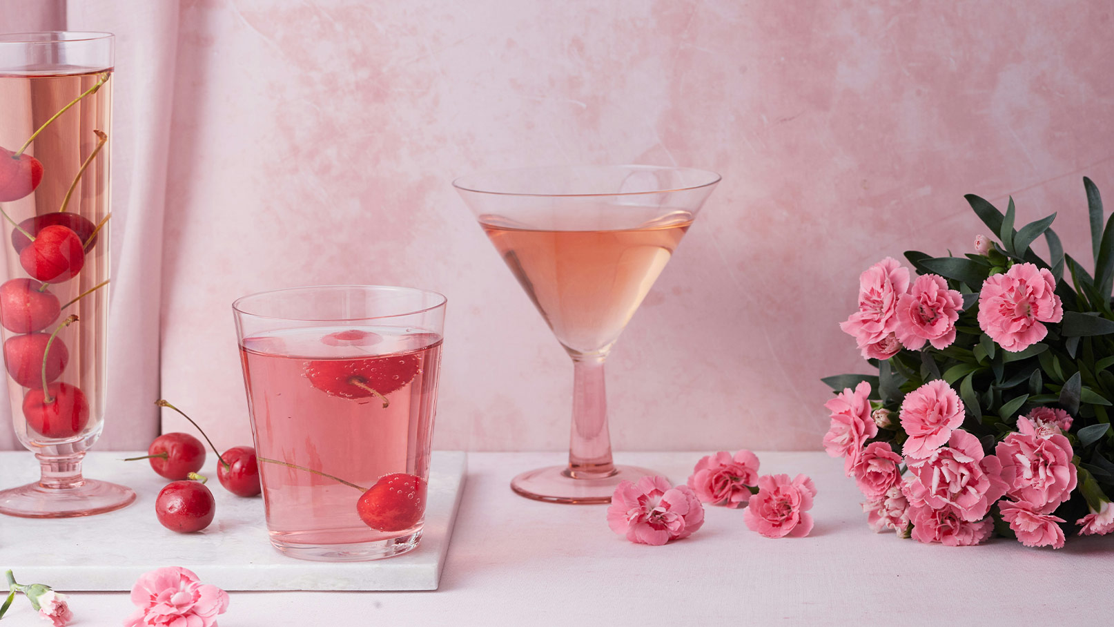Drei Gläser mit rosafarbenen Getränken sind vor einem sanften, rosa Hintergrund angeordnet. Eines der Gläser enthält Kirschen. Neben den Gläsern liegen pinke Blumen, die die fröhliche und einladende Stimmung der Szene unterstreichen.