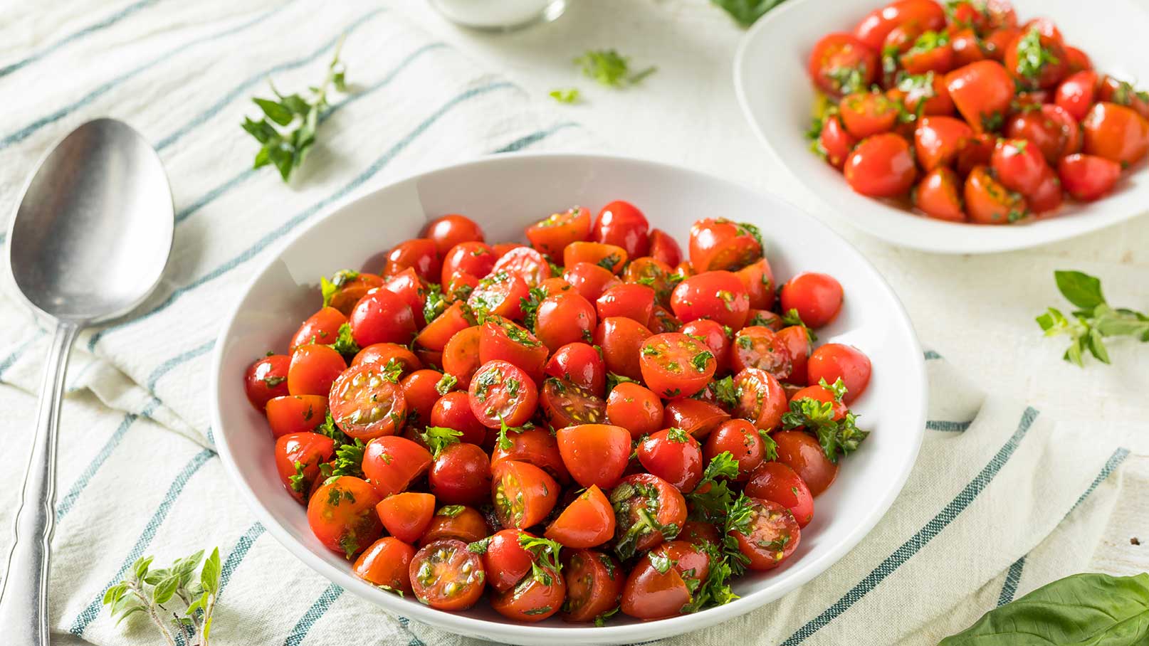 Eine große Schüssel gefüllt mit roten Cherrytomaten, gewürzt mit frischem, gehacktem Koriander. Die Tomaten sind halbiert und leuchten frisch. Im Hintergrund steht eine weitere Schüssel mit ähnlichen Tomaten. Ein Löffel und einige Kräuter liegen daneben auf einem gestreiften Tuch.
