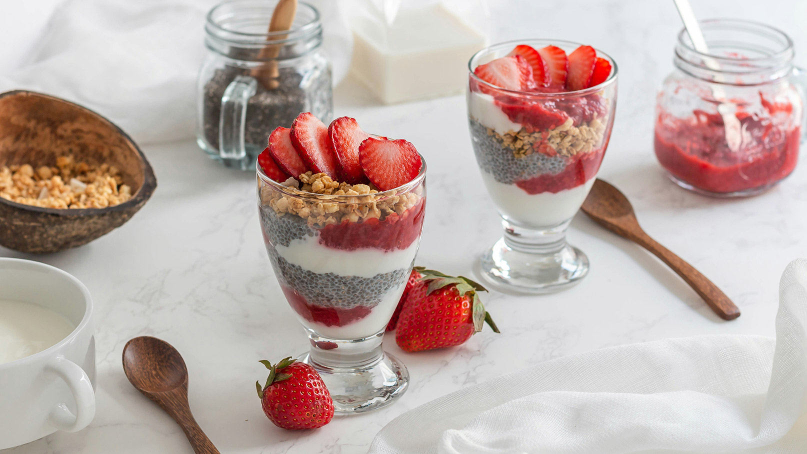 Zwei Gläser mit Schichtdessert sind auf einem weißen Tisch platziert. Jedes Glas enthält Joghurt, Chiasamen, Erdbeermarmelade, Hafergranola und frische Erdbeerscheiben obenauf. Daneben liegen ein kleiner Löffel und einige frische Erdbeeren.
