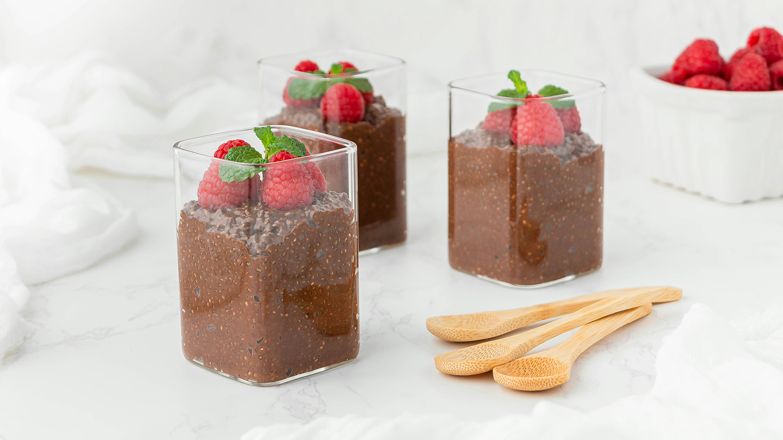 Drei Gläser mit Schokoladen-Chia-Pudding sind auf einem Tisch angerichtet, dekoriert mit frischen Himbeeren und Minzblättern. Daneben liegen drei kleine Holz-Löffel. Der Hintergrund ist hell und schlicht, um die Desserts hervorzuheben.