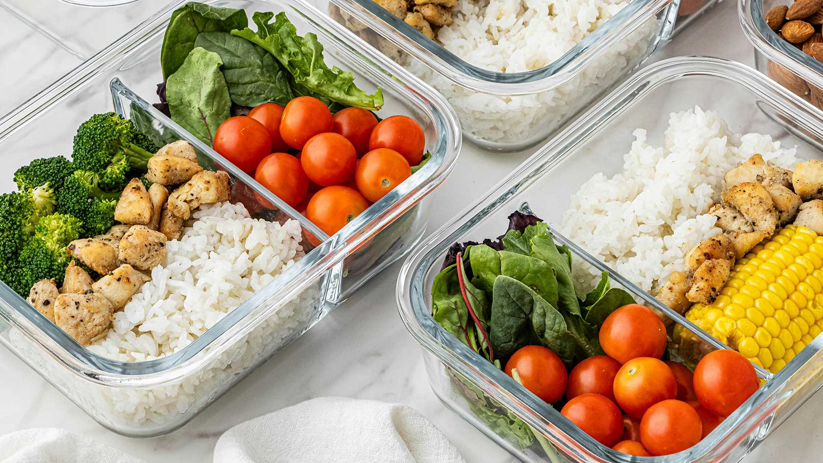 Drei transparente Behälter sind mit verschiedenen Lebensmitteln gefüllt. In einem Behälter befinden sich Brokkoli, gewürztes Hähnchen und Reis. Ein weiterer enthält Kirschtomaten, Spinat und Salat. Der dritte Behälter hat Reis, Hähnchenstücke und Mais. Die Speisen sind frisch zubereitet und ansprechend angerichtet.