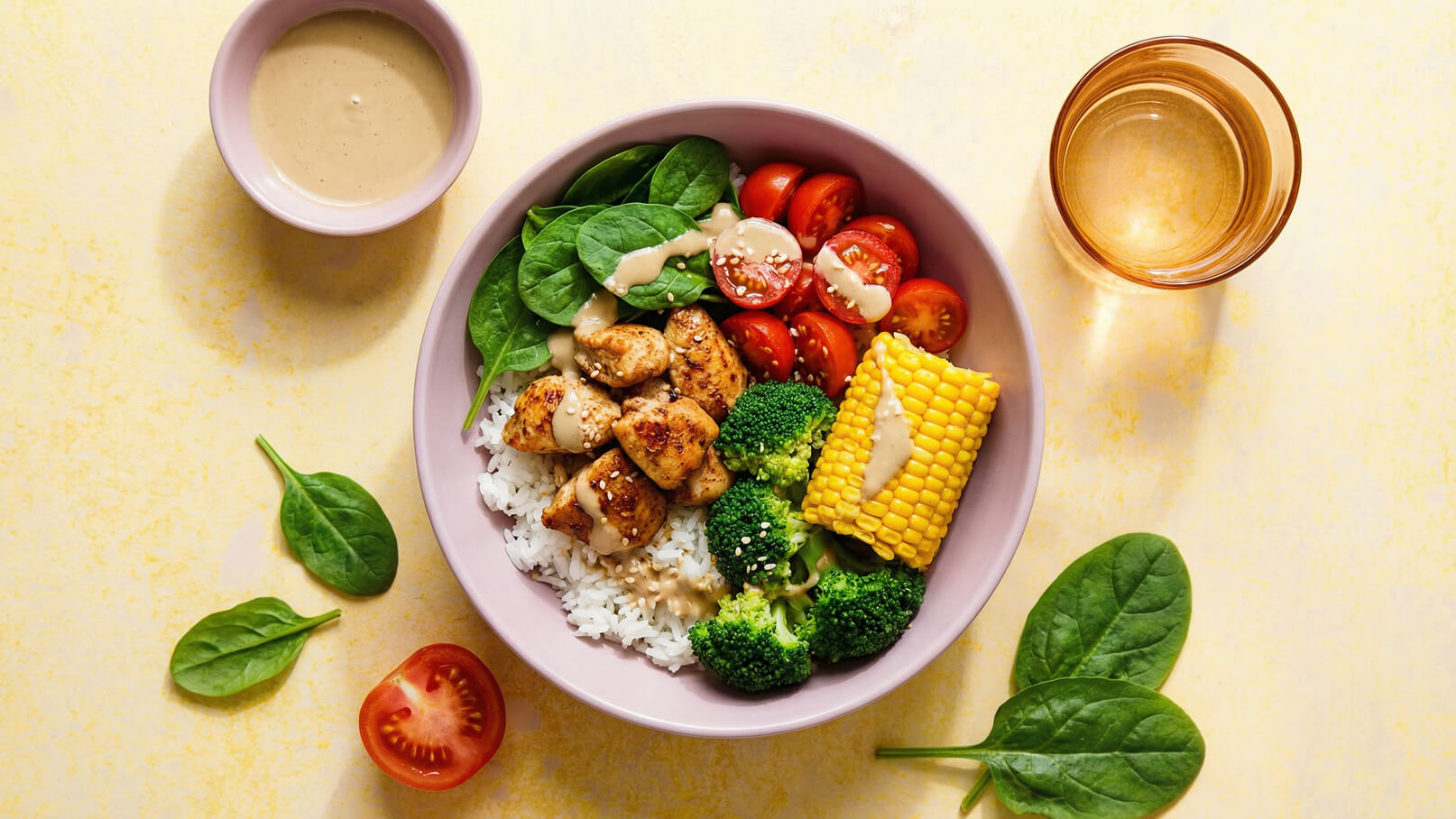Eine Schüssel mit Reis, gebratenem Hähnchen, frischem Spinat, Cherrytomaten, Brokkoli und einer Maiskolbenhälfte. Die Speisen sind mit einer cremigen Soße und Sesam bestreut. Daneben steht ein kleines Schälchen mit Soße und ein Glas Wasser. Der Hintergrund ist gelb.
