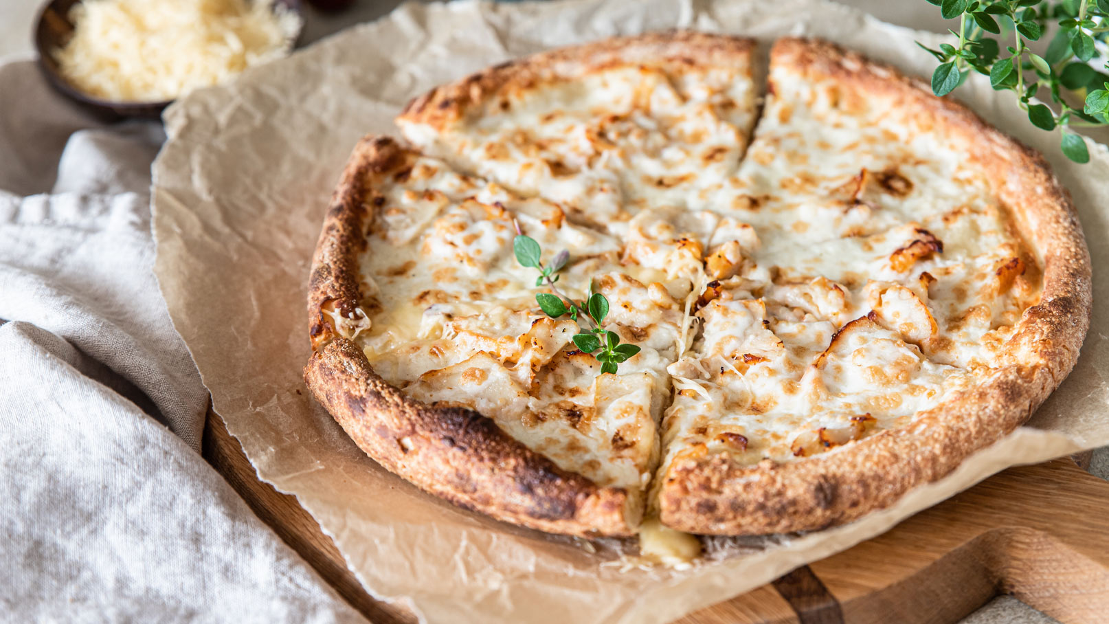 Eine goldbraune Pizza mit Käse und einem kleinen Zweig Oregano in der Mitte. Der Rand ist knusprig, und der Käse zeigt leichte Röststellen. Die Pizza liegt auf einem Backpapier und ist auf einem Holzbrett angerichtet.