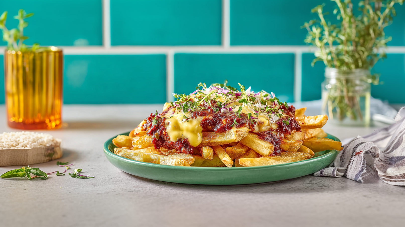 Ein grüner Teller mit Pommes Frites, bedeckt mit einer dicken Schicht Hackfleischsoße, geschmolzenem Käse und frischen Kräutern. Im Hintergrund ist ein Glas orangefarbener Saft und eine kleine Vase mit Kräutern sichtbar. Die Farben sind lebhaft und der Teller wirkt einladend.