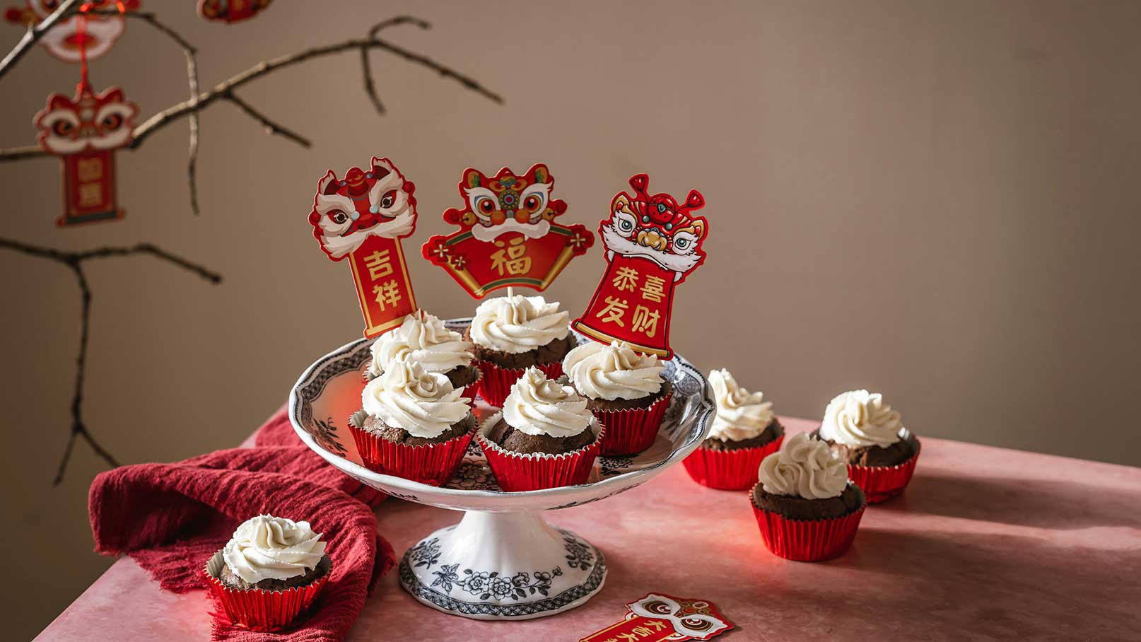 Ein Teller mit Schokoladen-Cupcakes, dekoriert mit weißer Sahne, steht auf einem Tisch. Einige Cupcakes sind rot umrandet, und es sind festliche Aufsteller mit Glückssymbolen in Rot und Gold zu sehen. Ein rotes Tuch liegt daneben.
