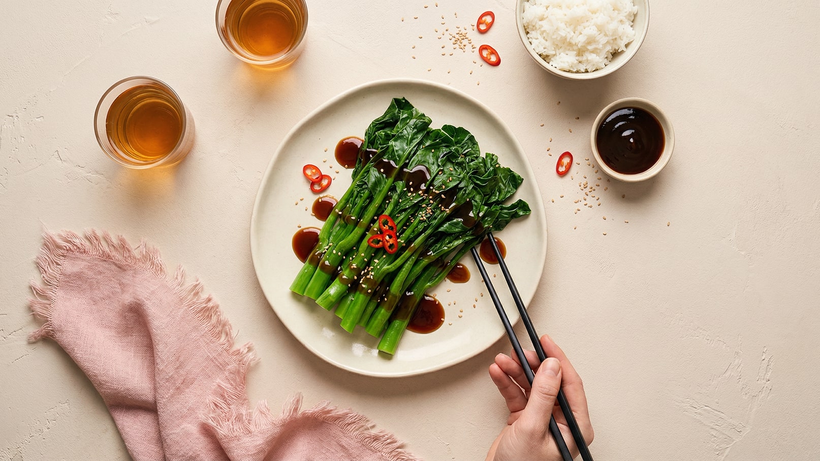 Ein Teller mit gekochtem Gemüse, garniert mit roten Chilis und Sesam, liegt auf einem Tisch. Eine Hand hält Essstäbchen, bereit, das Gemüse zu greifen. Im Hintergrund sind Gläser mit Tee und eine Schüssel Reis sichtbar.