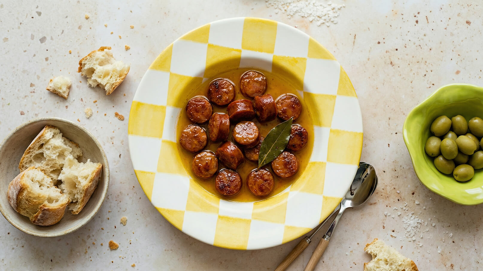 Ein Teller mit kleinen, gebratenen Würstchen in einer würzigen Sauce, garniert mit einem Lorbeerblatt. Neben dem Teller liegen Essbesteck und kleine Brötchenstücke, sowie eine Schüssel mit grünen Oliven. Der Hintergrund ist neutral und leicht strukturiert.