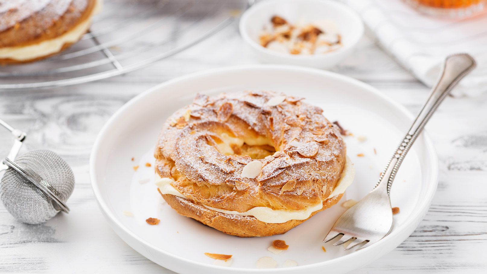 Ein frisch gebackener, runder Kuchen mit einer Füllung aus Sahne, bedeckt mit Puderzucker und gehobelten Mandeln. Der Kuchen liegt auf einem weißen Teller, neben einer kleinen Gabel. Im Hintergrund sind einige zerbrochene Mandelstücke sichtbar.