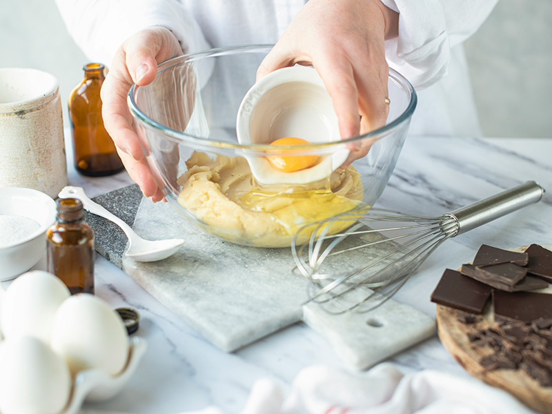 Eine Hand hält eine kleine Schüssel mit einem Ei über einer transparenten Schüssel. In der Schüssel liegt bereits Teig. Daneben stehen verschiedene Zutaten wie Zucker, Schokolade und Eier auf einer Arbeitsfläche. Ein Schneebesen liegt ebenfalls bereit.