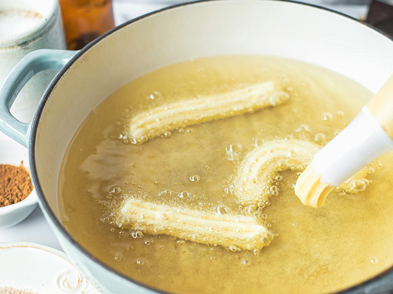 In einem großen Topf mit heißem Öl werden mehrere Churros frittiert. Sie sind goldbraun und schwimmen in der sprudelnden Flüssigkeit. Die Churros haben eine längliche Form und glänzen durch das Öl.