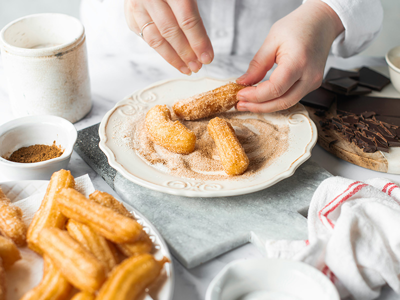 Eine Person bestreut frisch zubereitete Churros mit Zucker und Zimt, während sie einige auf einem Teller arrangiert. Im Hintergrund sind weitere Churros und Schüsseln mit Zutaten sichtbar.