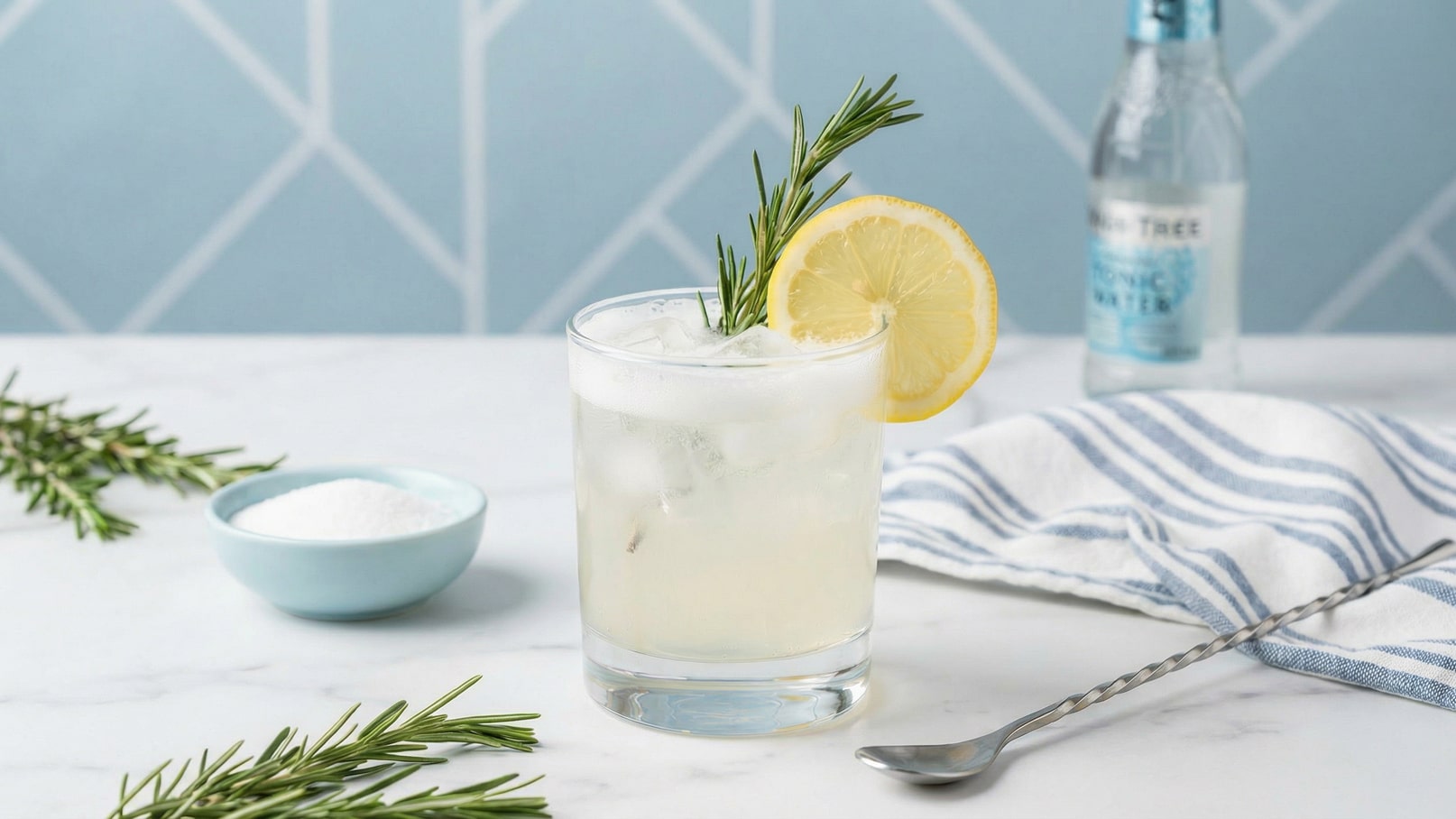 Ein Cocktailglas mit einem erfrischenden Getränk, das Eiswürfel, eine Zitronenscheibe und einen Rosmarinzweig enthält. Im Hintergrund befindet sich eine Flasche Tonic Water und eine Schüssel mit Zucker. Der Tisch hat eine helle, marmorierte Oberfläche.