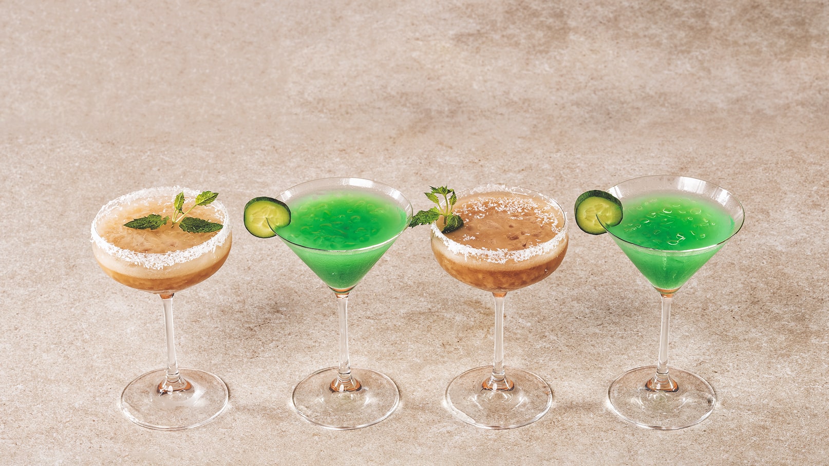 Drei Cocktails stehen in eleganten Gläsern auf einer neutralen Fläche. Der erste ist braun mit einer Zuckerrand und Minze, der zweite ist grün mit einem Limettenstück und der dritte kombiniert grünes und braunes Getränk, ebenfalls mit Minze.