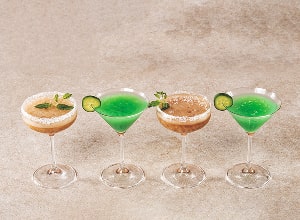 Vier Cocktails in eleganten Gläsern stehen nebeneinander. Die Getränke haben verschiedene Farben: ein Karotten-Cocktail, zwei grüne Cocktails und ein brauner Cocktail, die mit Gurkenscheiben und frischen Kräutern dekoriert sind. Der Hintergrund ist neutral und hebt die Drinks hervor.