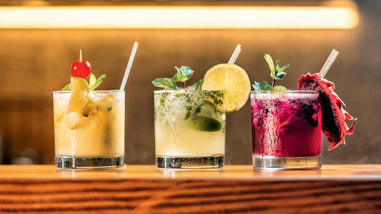 Drei Cocktails stehen auf einer Holzoberfläche. Der links ist fruchtig mit einer Kirsche und Minze. Der mittlere hat frische Limette und Minze. Der rechte Cocktail ist rot und enthält eine Drachenfrucht. Alle drei Getränke haben einen durchsichtigen Becher und sind mit einem Strohhalm versehen.