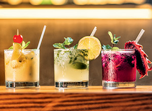 Drei Cocktails stehen auf einer Holzoberfläche: links ein gelber Cocktail mit Kirsche, in der Mitte ein grüner Mojito mit Limettenscheibe und rechts ein roter Drink mit Hibiskusblüten. Alle Cocktails haben Eiswürfel und einen Trinkhalm.