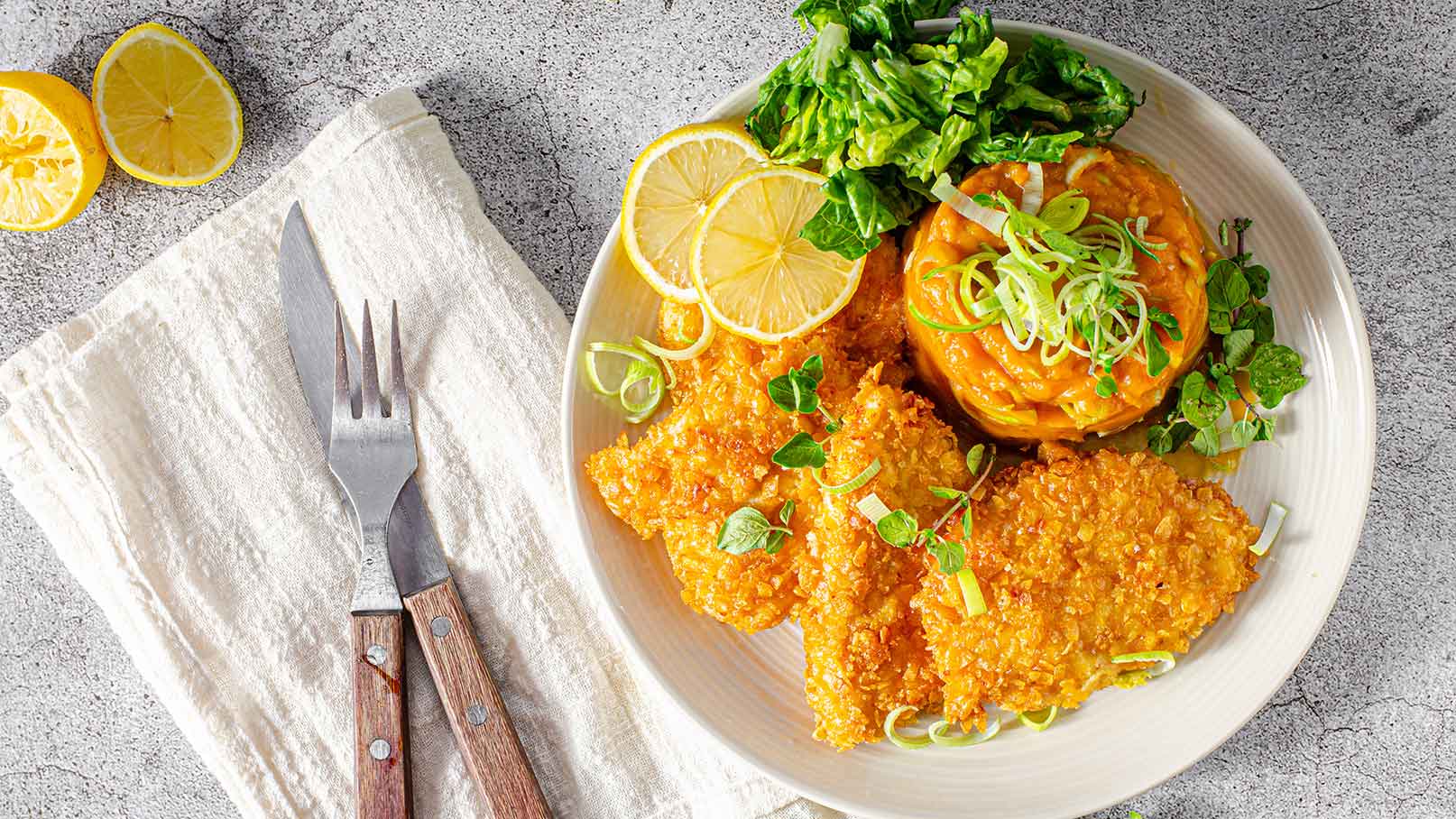Ein Teller mit knusprig gebratenem Fischfilet, umgeben von einem bunten Gemüse-Püree und frischem Salat. Dekoriert mit frischen Kräutern und Zitronenscheiben. Neben dem Teller liegen eine Gabel und ein Messer auf einer serviette.