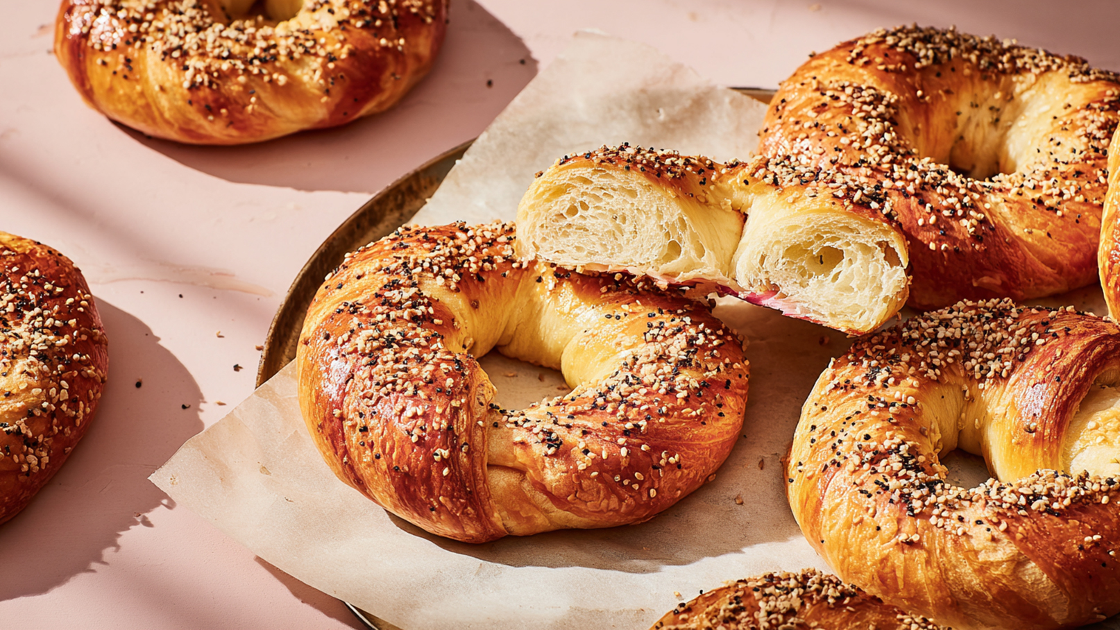 Eine Auswahl frisch gebackener Bagels, goldbraun und mit einer Mischung aus Sesam- und Schwarzkümmelsamen bestreut. Einer der Bagels ist zur Hälfte geschnitten, um die luftige, weiche Textur im Inneren zu zeigen. Hintergrund in sanften, neutralen Farbtönen.