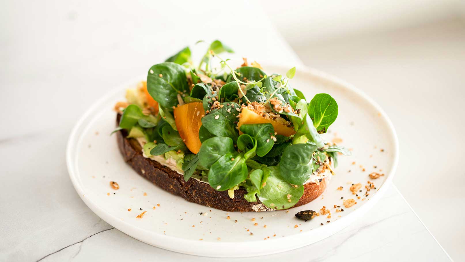 Ein Stück Vollkornbrot mit einer üppigen Salatmischung aus frischen, grünen Blättern und orangefarbenen Stückchen, verziert mit gerösteten Kernen. Der Teller hat eine glatte Oberfläche, und der Fokus liegt auf der bunten und gesunden Zusammenstellung.