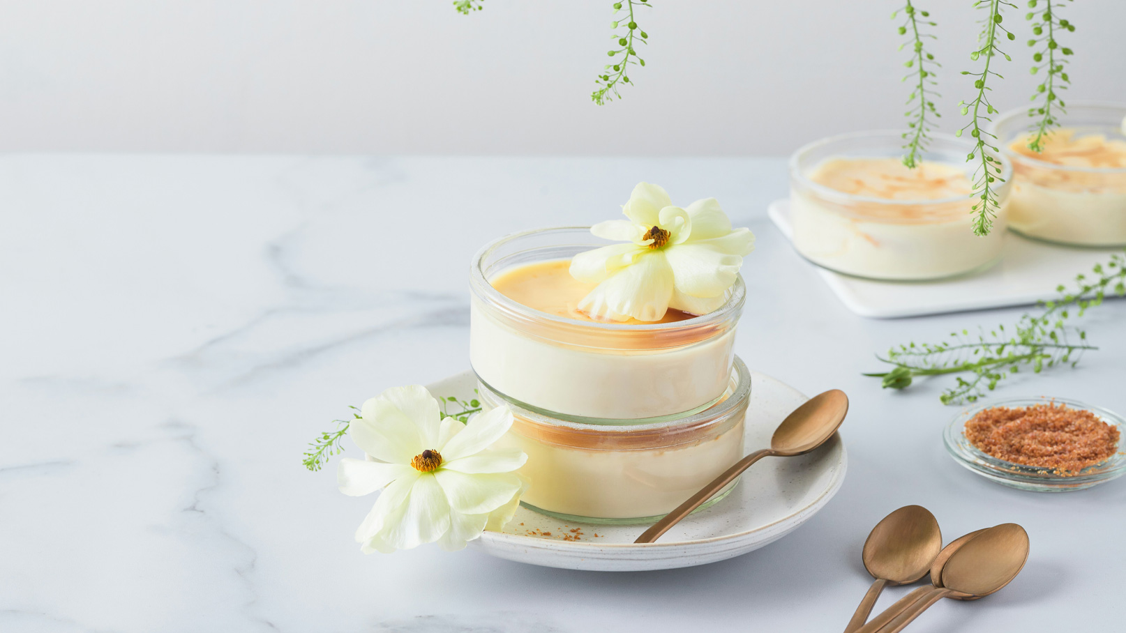 Zwei Gläser mit einem cremigen Dessert stehen auf einem Teller. Eines der Gläser ist höher. Beide sind mit zarten, weißen Blumen dekoriert. Im Hintergrund sind weitere Dessertgläser und ein kleiner Teller mit braunem Zucker sowie Löffel zu sehen.