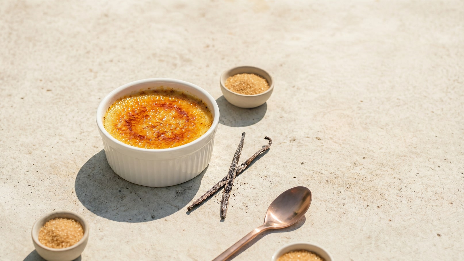 Eine Schale mit crème brûlée steht auf einer hellen Oberfläche. Die Creme hat eine goldene, karamellisierte Oberfläche. Neben der Schale sind zwei Vanilleschoten und kleine Schalen mit Zucker. Löffel aus Metall liegen ebenfalls daneben.