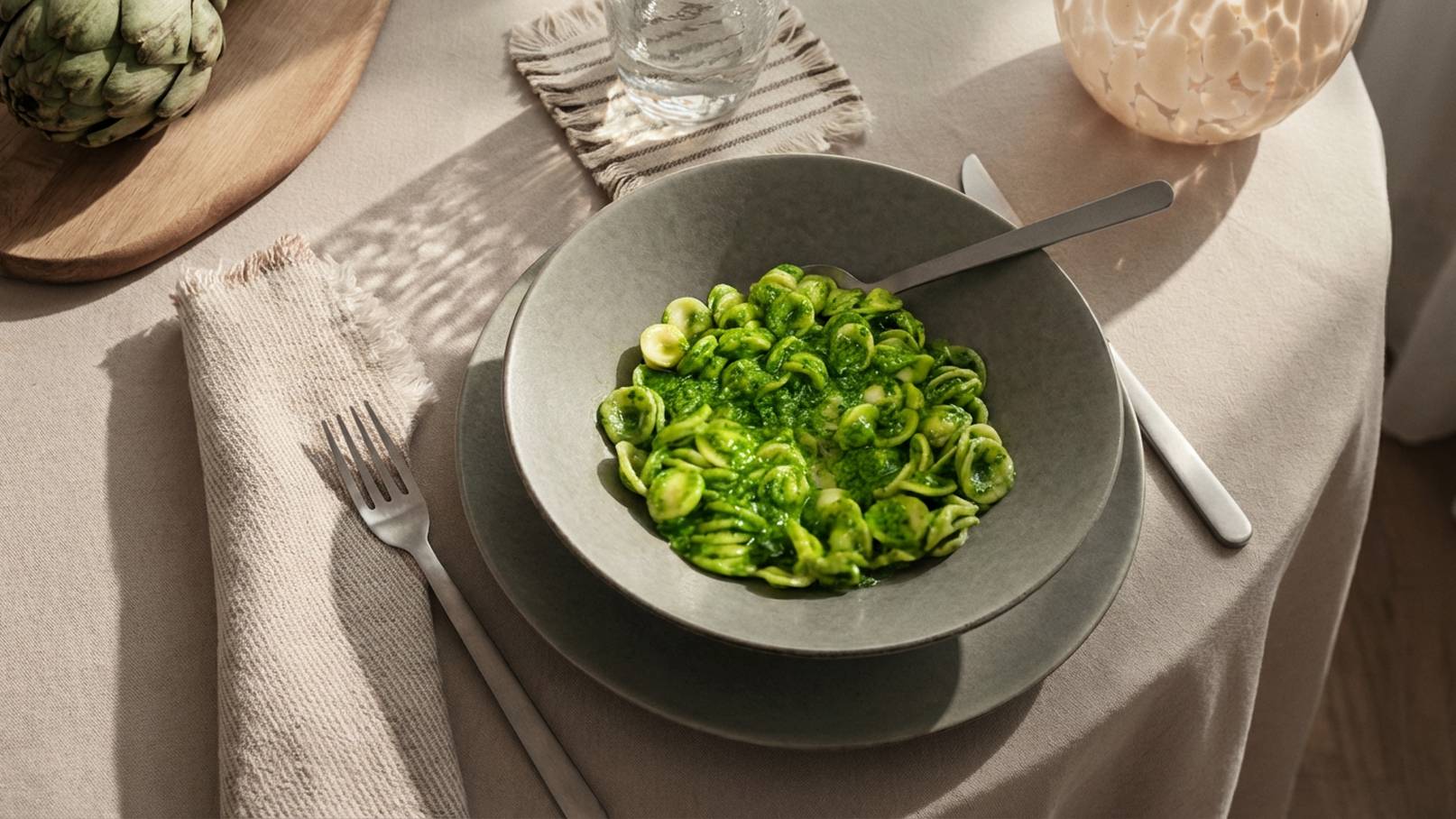 Ein grauer Teller mit grünem Pasta-Gericht, präsentiert auf einem gedeckten Tisch. Dazu gehören Besteck, eine Wasserflasche und einweiches Tischtuch. Die helle Beleuchtung betont die frischen Farben der Speise.