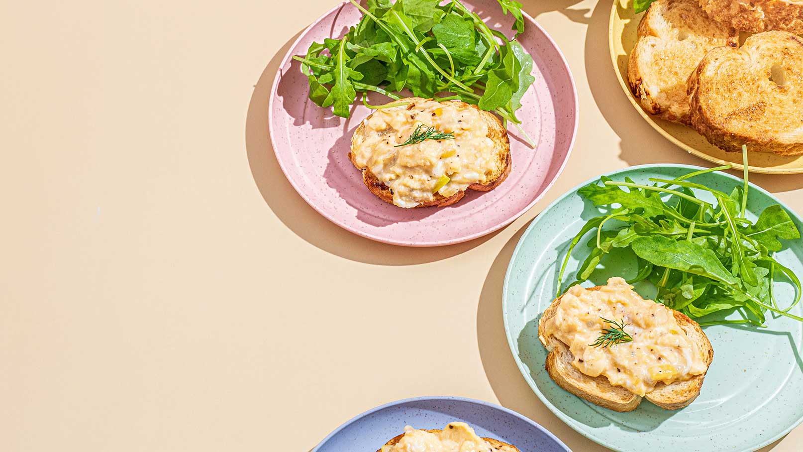 Auf den Tellern befinden sich mehrere Portionen eines herzhaften Gerichts, begleitet von frischem Rucola. Die Teller sind in verschiedenen Farben (rosa, blau und gold) angeordnet, und im Hintergrund sind einige getoastete Brotscheiben sichtbar.