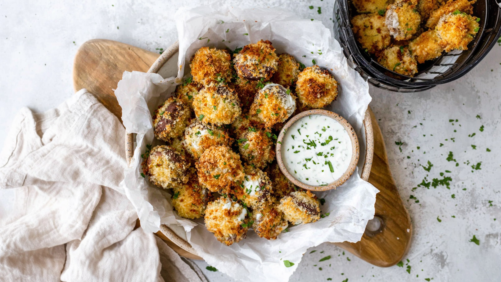 Eine Schüssel mit knusprig gebratenen Bällchen, umgeben von Pergamentpapier. Neben den Bällchen steht eine kleine Schale mit weißer Dipsauce, garniert mit grünen Kräutern. Der Hintergrund ist schlicht und zeigt eine Holzplatte.