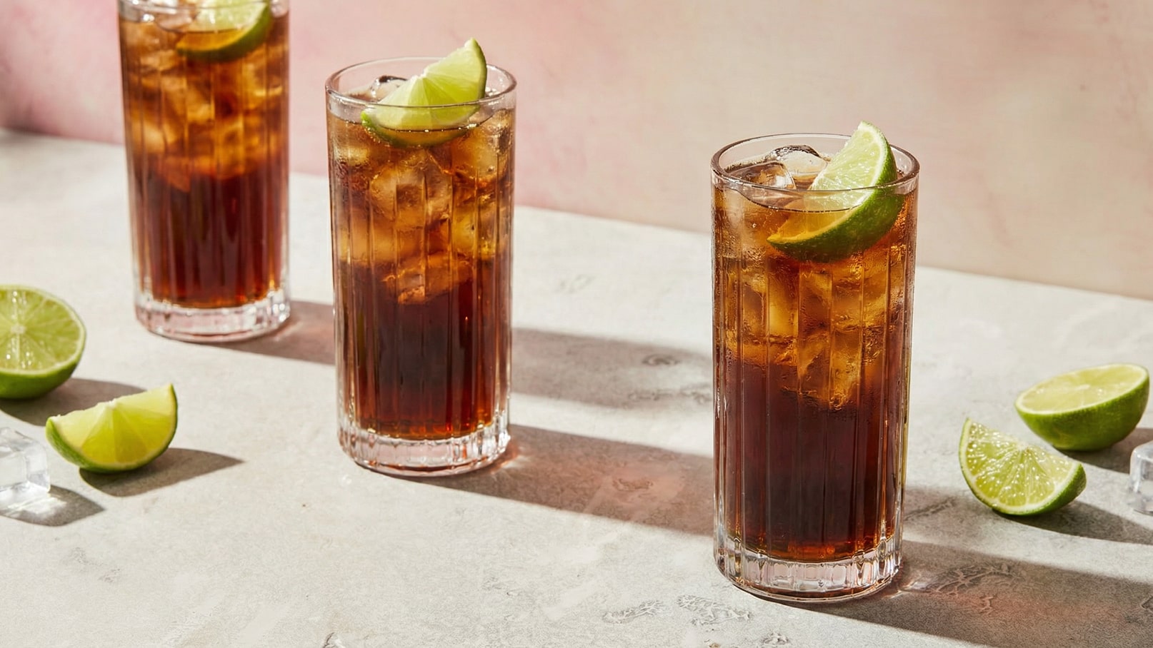 Drei gläserne Cocktails mit Eiswürfeln und Limettenscheiben, serviert auf einer hellen, strukturierten Oberfläche. Neben den Drinks liegen einige halbierte Limetten. Der Hintergrund ist sanft verschwommen und in Pastelltönen gehalten.