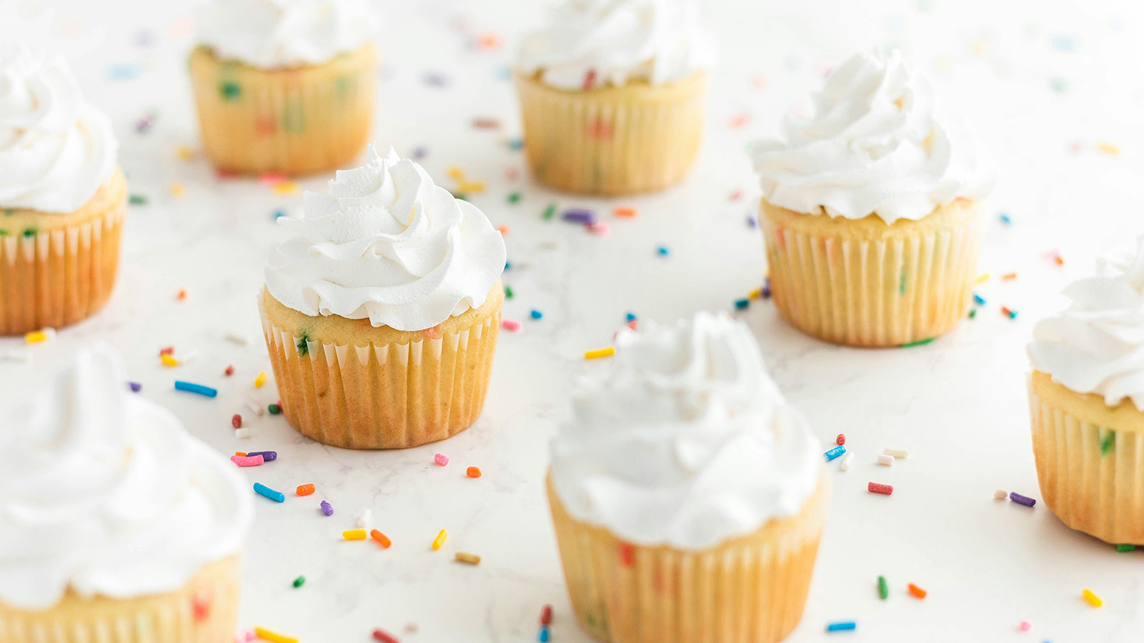 Der Bildinhalt zeigt mehrere farbenfrohe Cupcakes mit einer glänzenden, weißen Sahnehaube. Die Cupcakes sind auf einer hellen Oberfläche angeordnet und von bunten Streuseln umgeben. Sie wirken frisch und einladend.