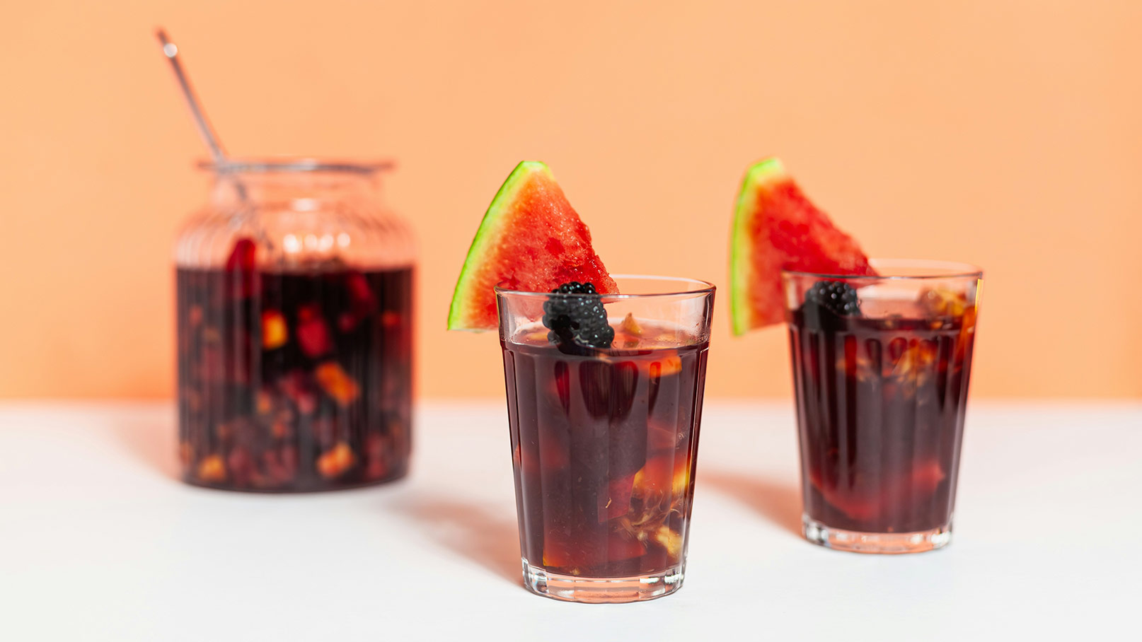 Zwei Gläser mit dunklem Getränk, garniert mit einem Stück Wassermelone und einer Brombeere. Im Hintergrund steht ein großer, ebenfalls gefüllter Glasbehälter mit dem gleichen Getränk. Der Hintergrund ist in sanften Pfirsichtönen gehalten.
