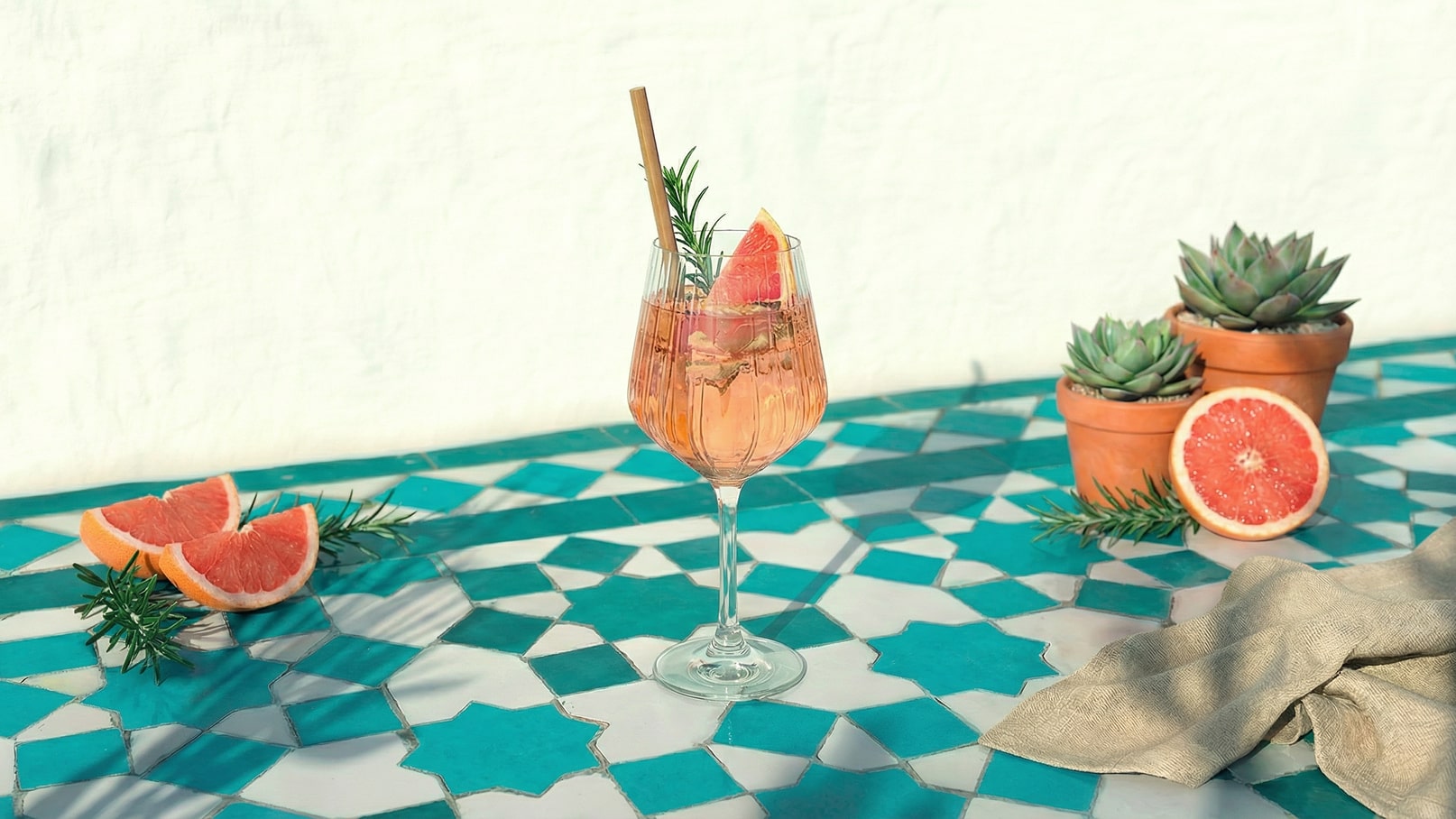 Ein elegantes Glas mit einem rosa Getränk steht auf einem bunten, geometrischen Tisch. Das Getränk ist mit einer Scheibe Grapefruit und frischen Kräutern garniert. Neben dem Glas liegen halbierte Grapefruits und einige kleine Pflanzentöpfe mit Sukkulenten.