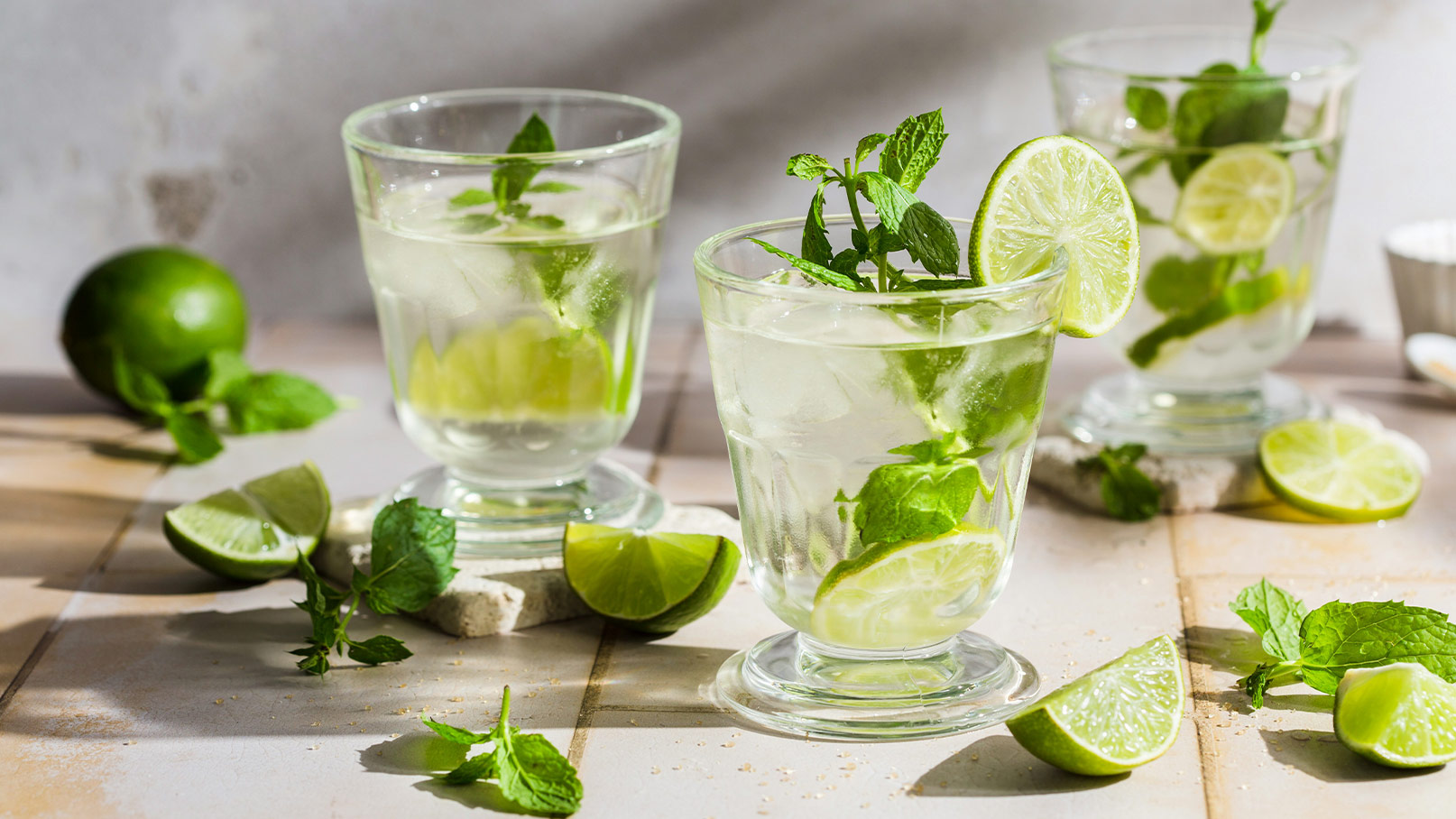 Drei Gläser mit einem erfrischenden Getränk, gefüllt mit Eis, Minzblättern und Limettenscheiben, stehen auf einem Holztisch. Rund um die Gläser liegen frische Limetten und Minzweigen. Das Bild vermittelt ein Gefühl von sommerlicher Frische und Genuss.
