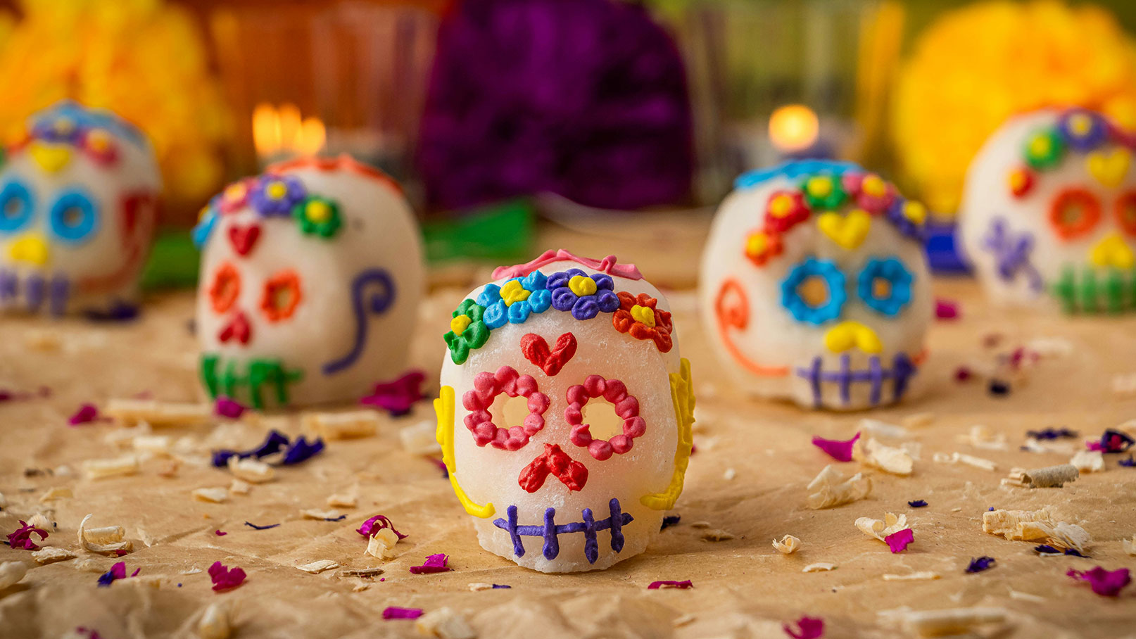 Das Bild zeigt bunte Zuckerschädel, die mit lebhaften Dekorationen wie Blumen und Herzen verziert sind. Sie sind Teil der traditionellen Feierlichkeiten zum Día de los Muertos. Der Hintergrund ist mit bunten Blumen und Kerzen geschmückt, die eine festliche Atmosphäre schaffen.