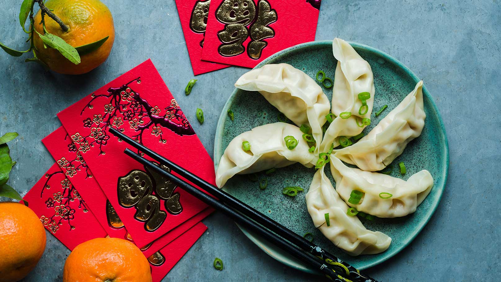 Ein Teller mit gefüllten Teigtaschen (Dumplings), die kreisförmig angeordnet sind, auf einem blauen Untergrund. Frisch geschnittene Frühlingszwiebeln liegen auf den Dumplings. Neben dem Teller befinden sich zwei Orangen und rote Umschläge mit goldenen Verzierungen.
