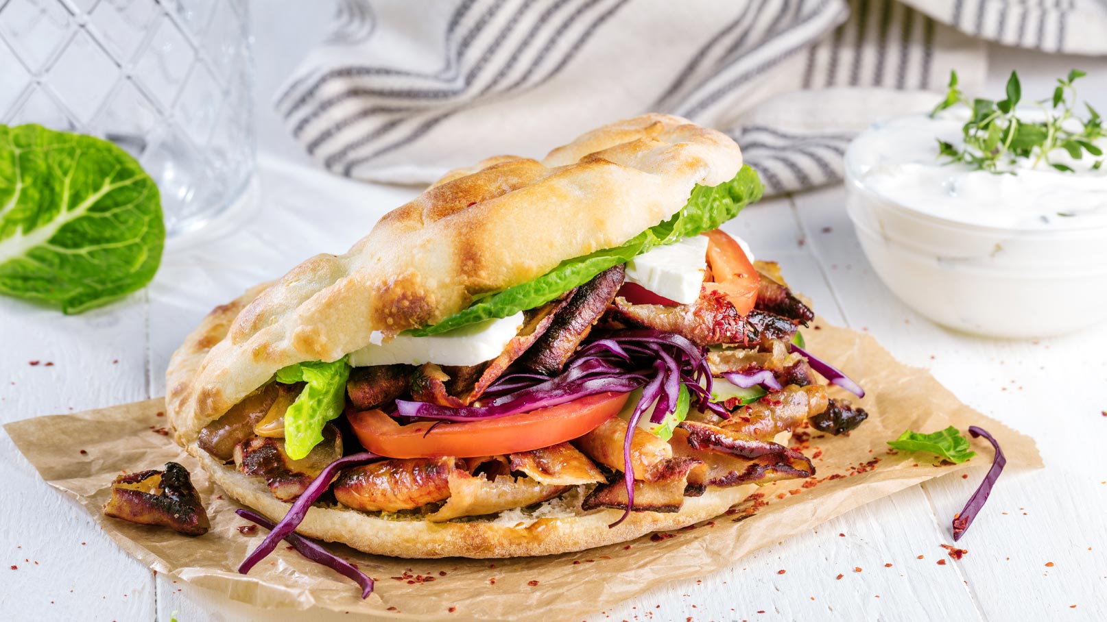 Ein frisch zubereiteter Sandwich mit knusprigem Brot, gefüllt mit gegrilltem Fleisch, roten Zwiebeln, Tomaten und Salat. Darauf liegen Scheiben von Feta-Käse. Das Sandwich liegt auf braunem Papier und wird von einem Becher mit Joghurtsoße begleitet.