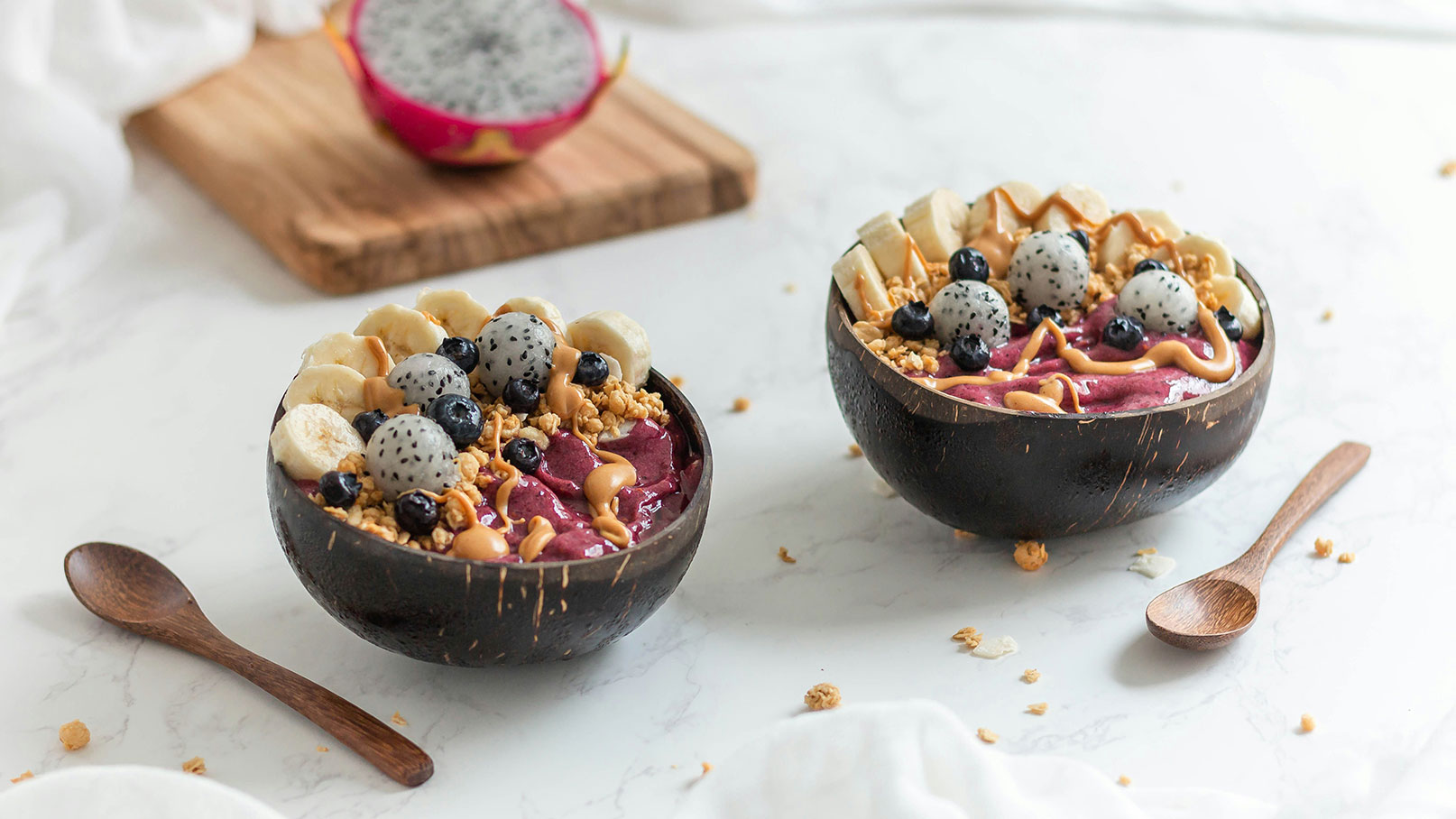 Zwei Schalen mit Smoothie-Bowl, gefüllt mit püriertem Obst, zerkleinerten Cashewkernen, Bananenstücken, Blaubeeren und grünem Drachenfrucht- dekoriert. Neben den Schalen liegt ein Holzlöffel. Die Schalen sind aus Kokosnuss. Der Hintergrund ist hell und schlicht.