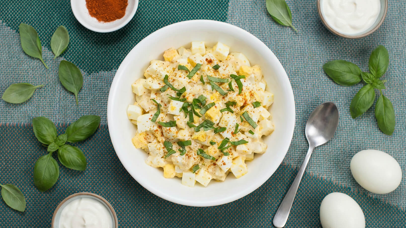 Eine weiße Schüssel mit einem Salat aus gewürfelten Eiern, Mayonnaise und frischem Basilikum. Der Salat ist mit Paprika und frischen Kräutern garniert. Auf dem Tisch liegen einige frische Basilikumblätter und eine kleine Schale mit Joghurt.