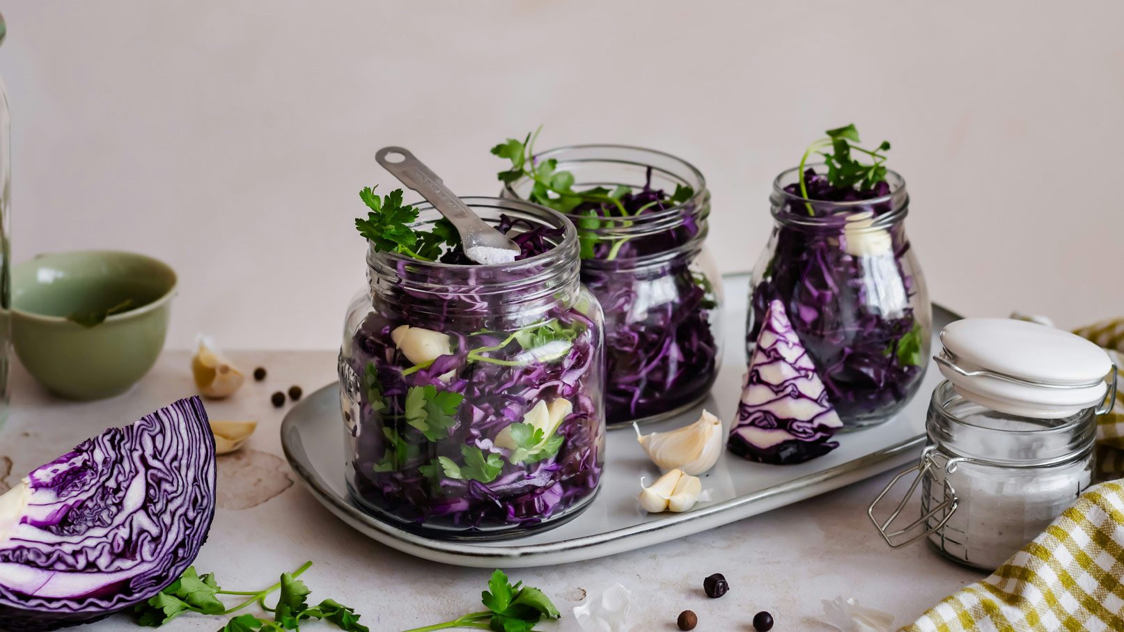 Drei Gläser mit eingelegtem Rotkohl, verziert mit frischem Koriander und Knoblauchzehen, stehen auf einem Tablett. Ein halber Kopf Rotkohl und Gewürze sind daneben platziert. Die Szene vermittelt eine frische und gesunde Küchenatmosphäre.