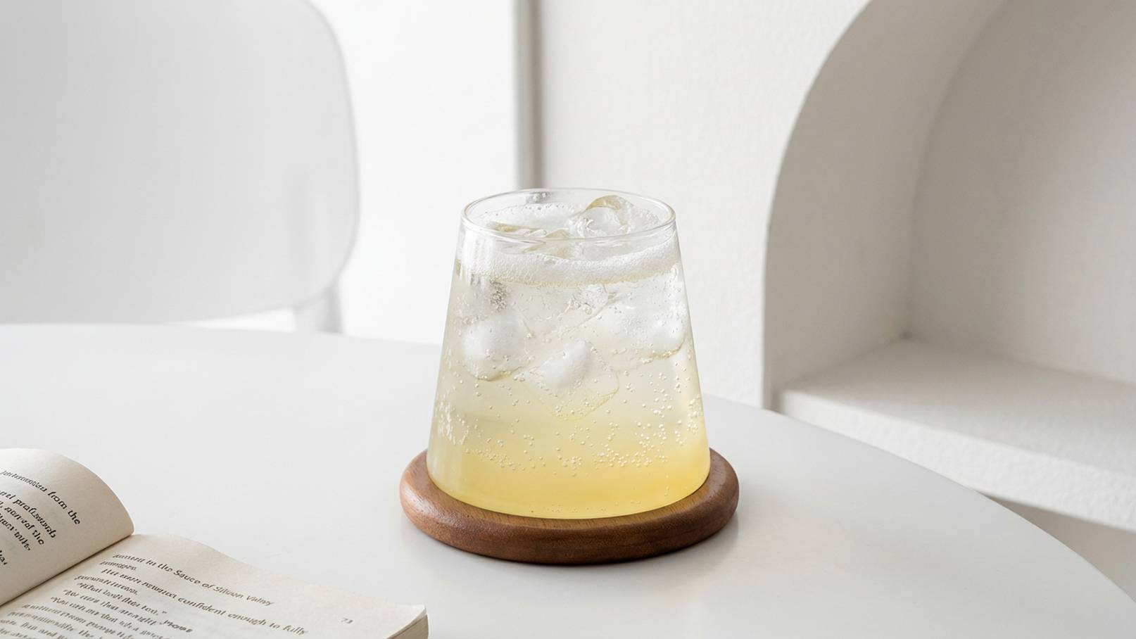 Ein Glas mit eisgekühltem Getränk und blubbernder Limonade steht auf einem Holzuntersetzer auf einem Tisch. Im Hintergrund liegt ein aufgeschlagenes Buch. Die Atmosphäre ist frisch und einladend.