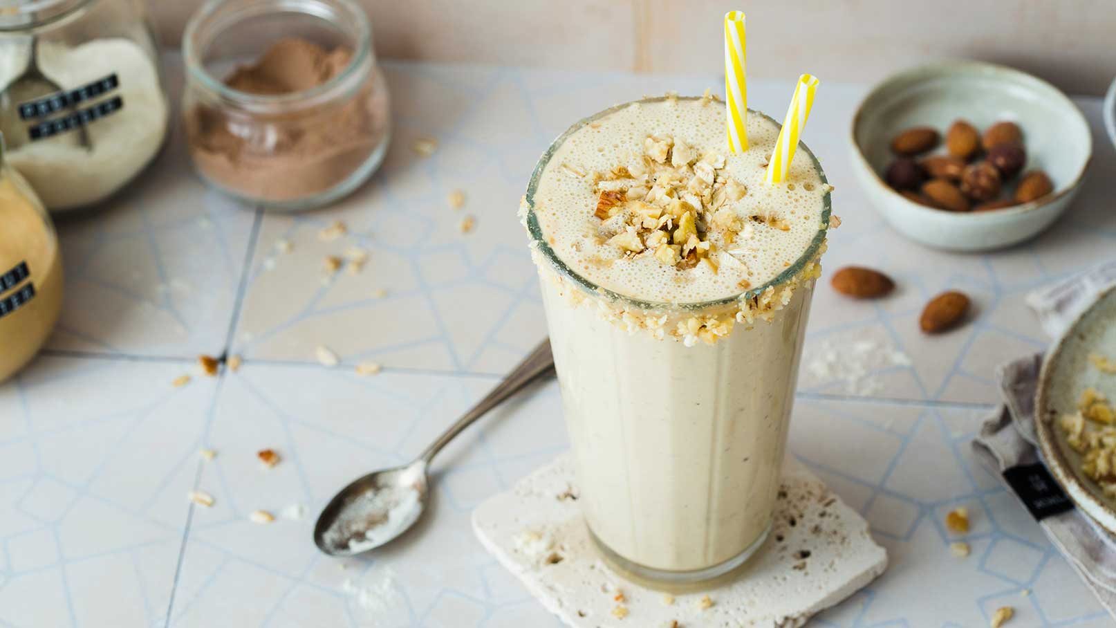 Ein hoher Glasbecher mit einem cremigen Shake, garniert mit gehackten Nüssen und zwei gelben Strohhalmen. Der Becher steht auf einem Untersetzer und ist umgeben von verschiedenen Zutaten, die auf einer hellen, marmorierten Oberfläche verteilt sind.