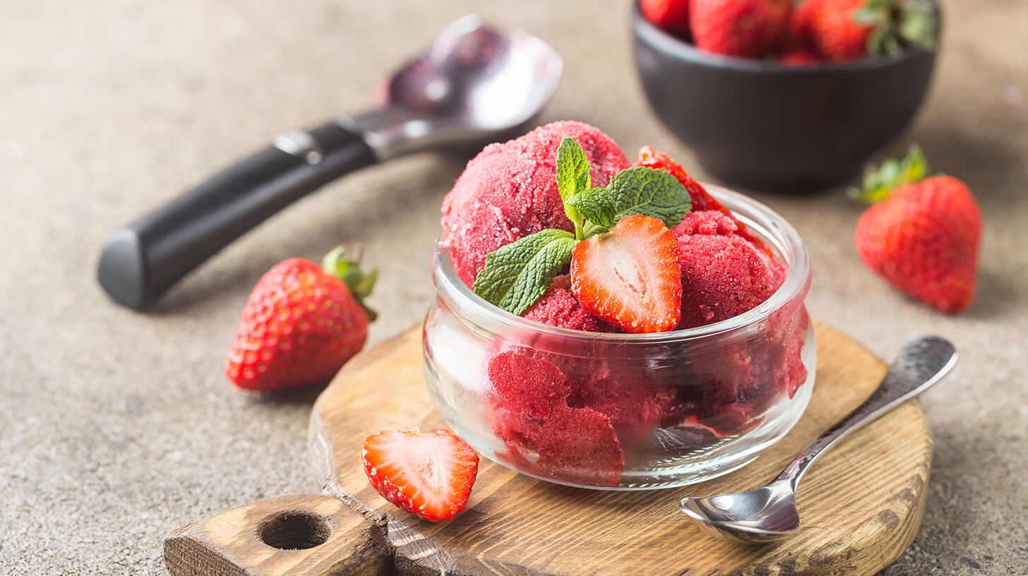 Ein Glas mit Erdbeersorbet, garniert mit frischen Erdbeerscheiben und einem Minzblatt, steht auf einem Holzbrett. Daneben liegt ein kleiner Löffel. Im Hintergrund sind weitere Erdbeeren und ein dunkles Schälchen zu sehen.