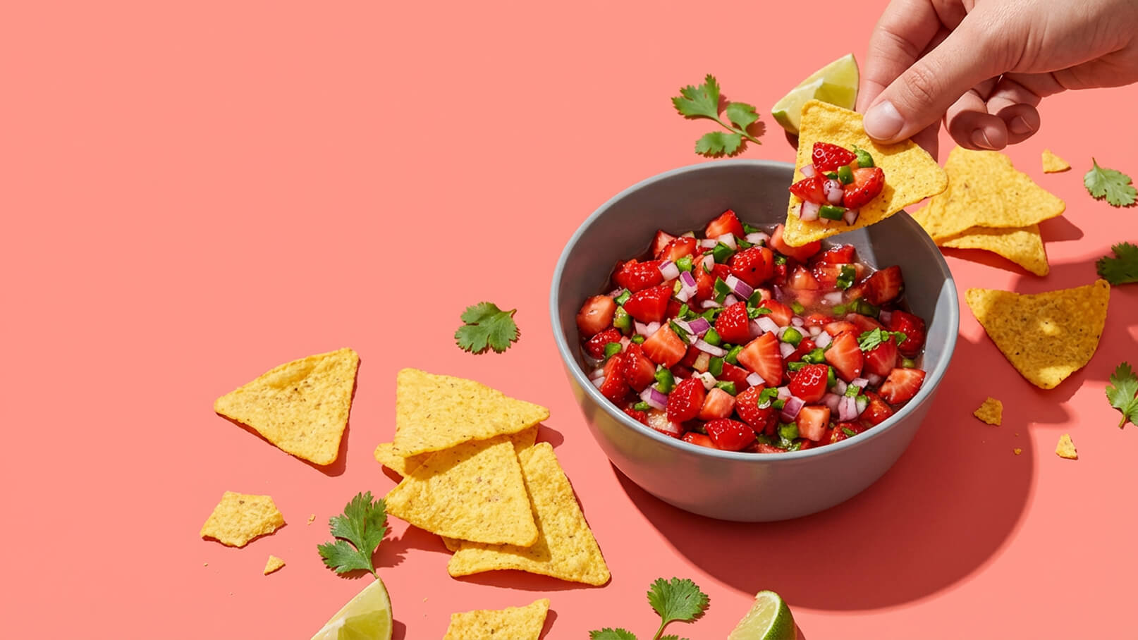 Eine Hand hält einen Tortilla-Chip über eine Schüssel mit frischer Salsa, die aus Erdbeeren, Zwiebeln und Koriander besteht. Rund um die Schüssel liegen einige weitere Tortilla-Chips und eine Limette. Der Hintergrund ist in einem sanften Rosa gehalten.