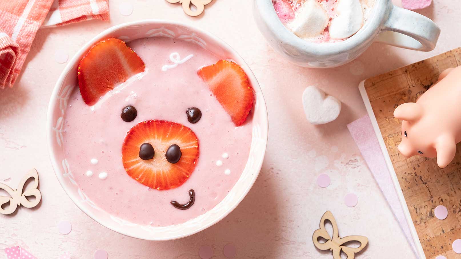 Eine Schüssel mit pinkem Joghurt in Form eines Schweins, dekoriert mit Erdbeeren als Ohren und Gesicht, sowie Schokoladenstückchen als Augen. Neben der Schüssel steht eine Tasse mit einer milchigen Getränkemischung und einigen Marshmallows.