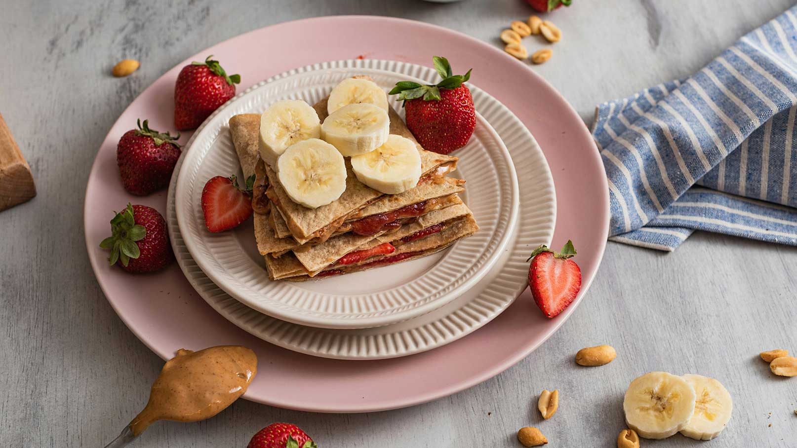 Ein Teller mit gestapelten, dünnen Brotscheiben, gefüllt mit Erdbeermarmelade und Bananenscheiben. Aromatische Erdbeeren sind um den Teller herum angeordnet, und ein Löffel Erdnussbutter liegt daneben. Die Anordnung vermittelt eine appetitliche und frische Snack-Idee.