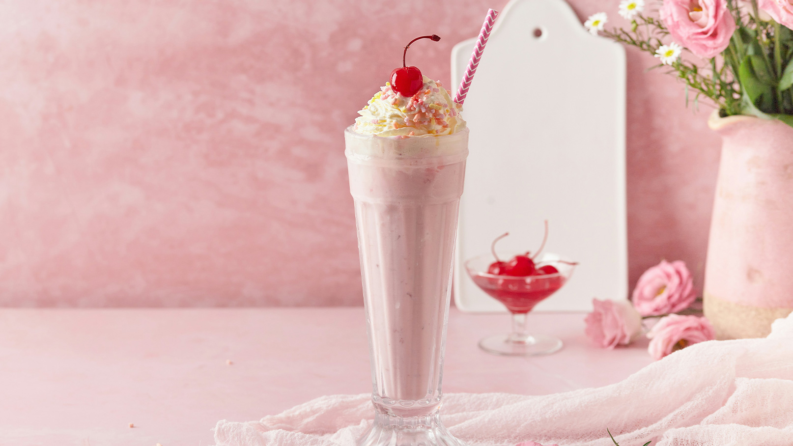 Ein köstlicher Milchshake in einem hohen Glas, garniert mit Sahne, bunten Streuseln und einer Kirsche obenauf. Im Hintergrund steht ein weiterer kleiner Dessertbecher mit Kirschen. Alles auf einem sanften, rosa Hintergrund.