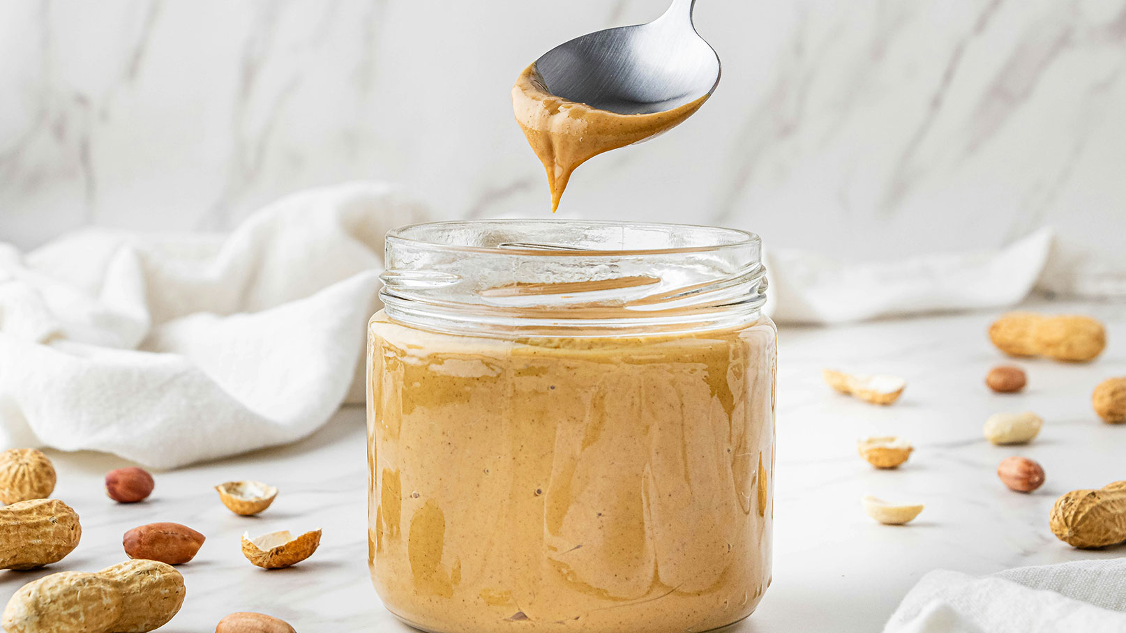 Ein Glas Erdnussbutter steht auf einem Tisch, während ein Löffel eine Portion Erdnussbutter darüber hält. Um das Glas herum liegen einige ungeschälte und geschälte Erdnüsse. Der Hintergrund ist hell und neutral, was die Erdnussbutter hervorhebt.