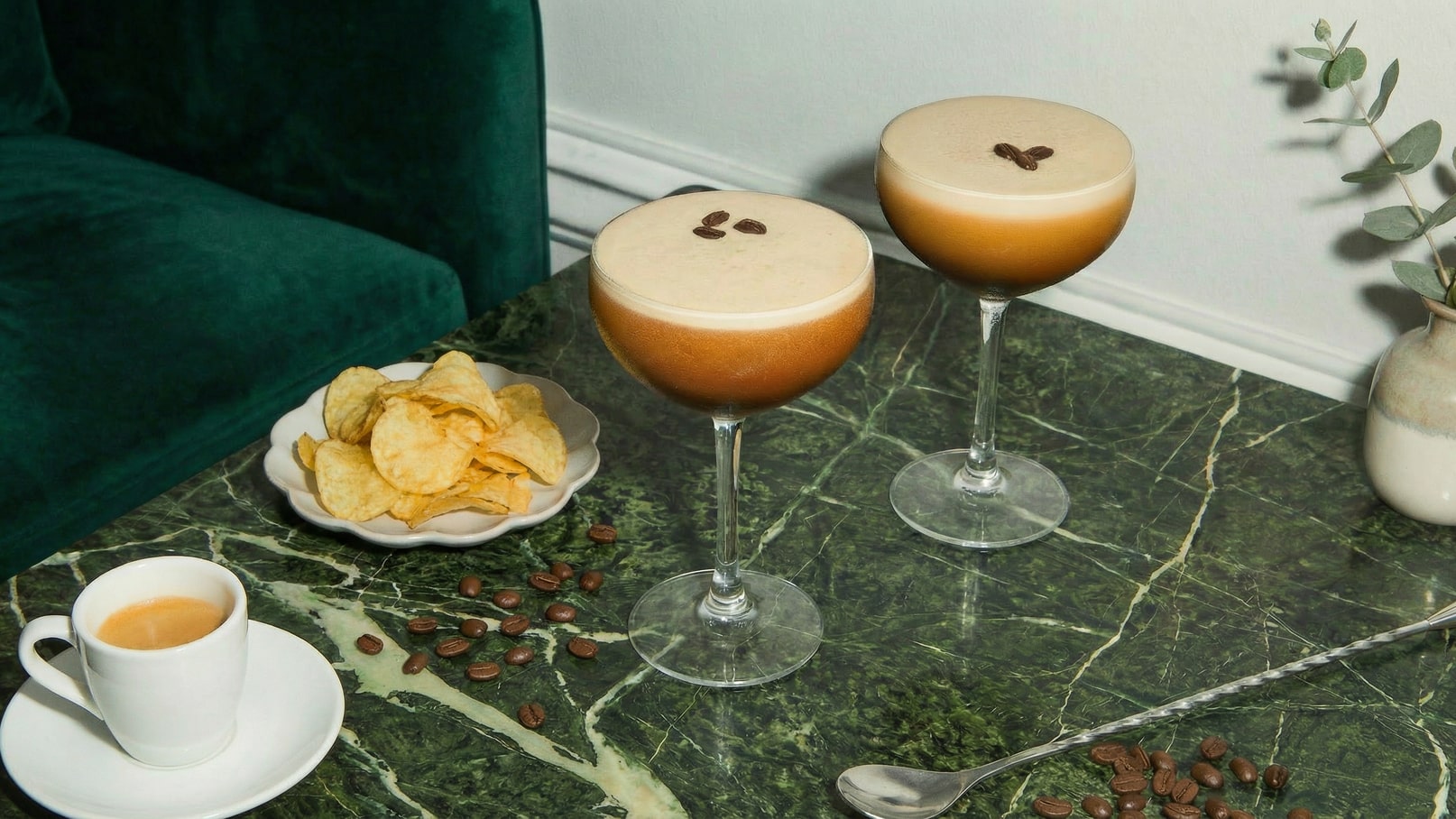 Zwei Cocktails in eleganten Gläsern stehen auf einem marmorierten Tisch, dekoriert mit Kaffeebohnen. Neben ihnen liegt ein Teller mit knusprigen Kartoffelchips. In der Nähe steht eine kleine Tasse Espresso.