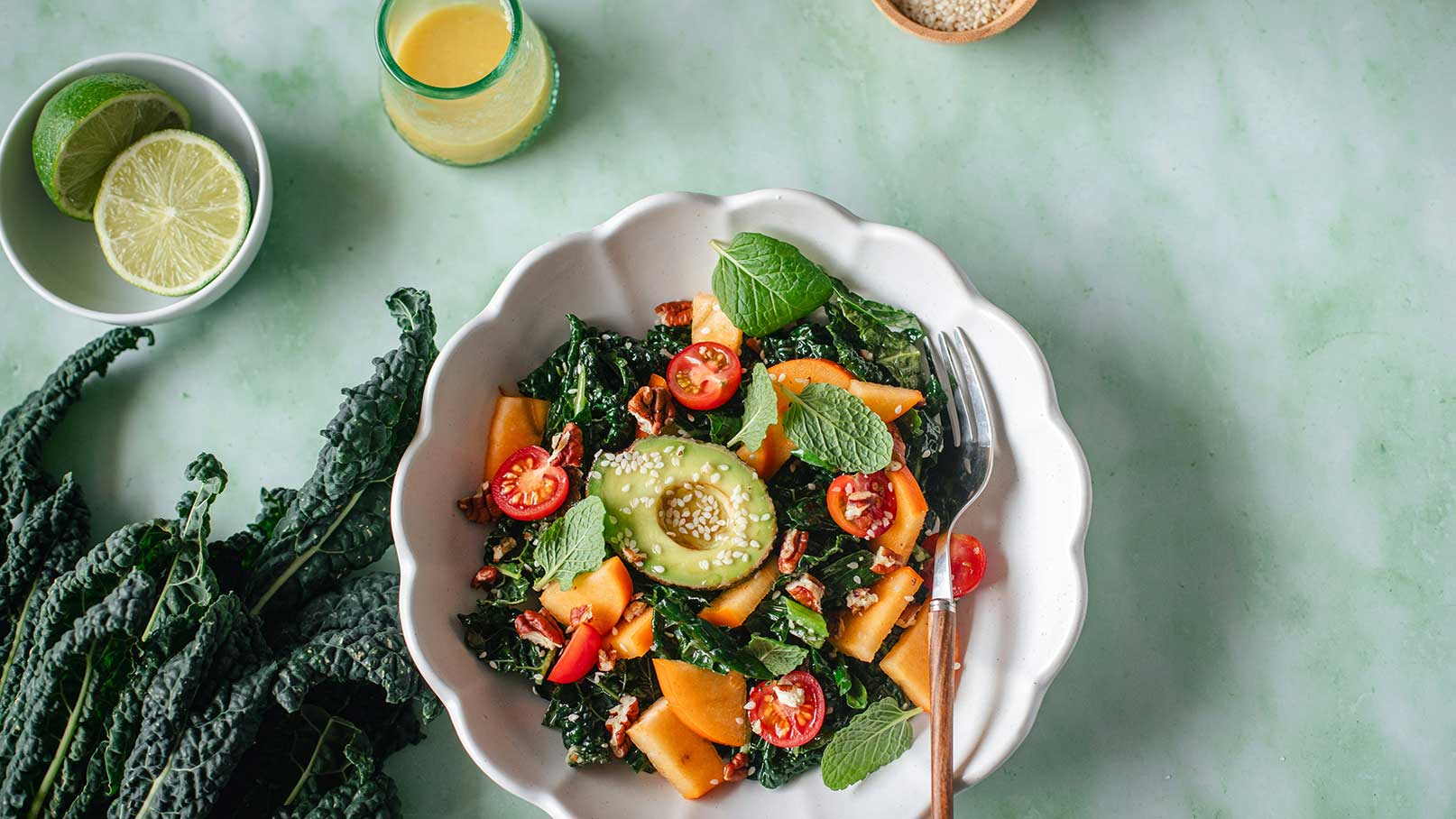 Eine Schüssel mit buntem Salat, bestehend aus frischem Blattgemüse, Kirschtomaten und orangefarbenen Stücken, möglicherweise von einer Melone oder einer ähnlichen Frucht. Ein Essbesteck liegt daneben, während einige Zutaten im Hintergrund sichtbar sind.